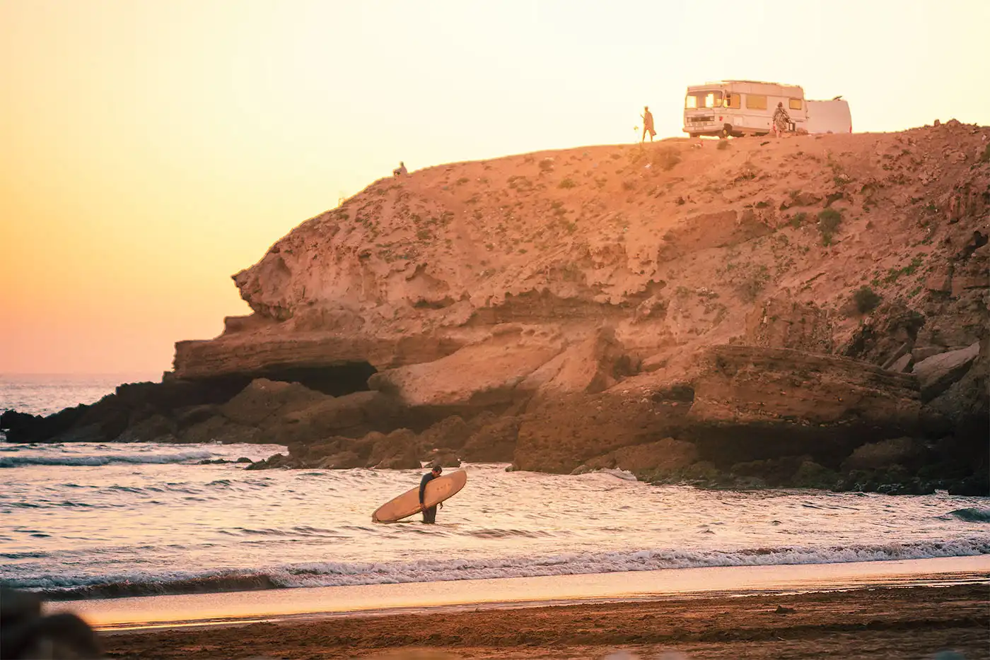 person walking to shore with surfboard at sunset